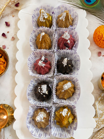 A white scalloped serving tray holding ten small paper cup liners filled with assorted sweets or confections, each topped with a sprinkle of powdered sugar. The tray is photographed from above on a light marble-like surface with decorative props around it, including a peacock feather, orange marigold, and small brass dishes.