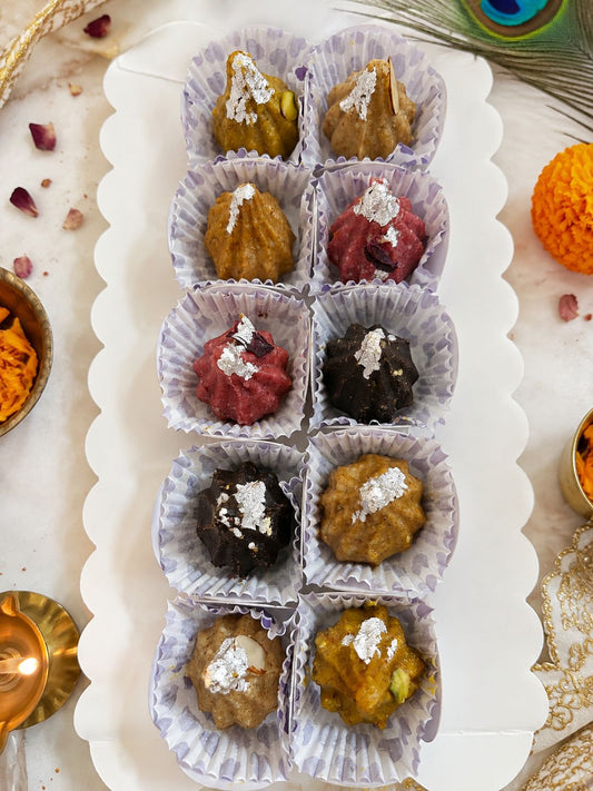 A white scalloped serving tray holding ten small paper cup liners filled with assorted sweets or confections, each topped with a sprinkle of powdered sugar. The tray is photographed from above on a light marble-like surface with decorative props around it, including a peacock feather, orange marigold, and small brass dishes.